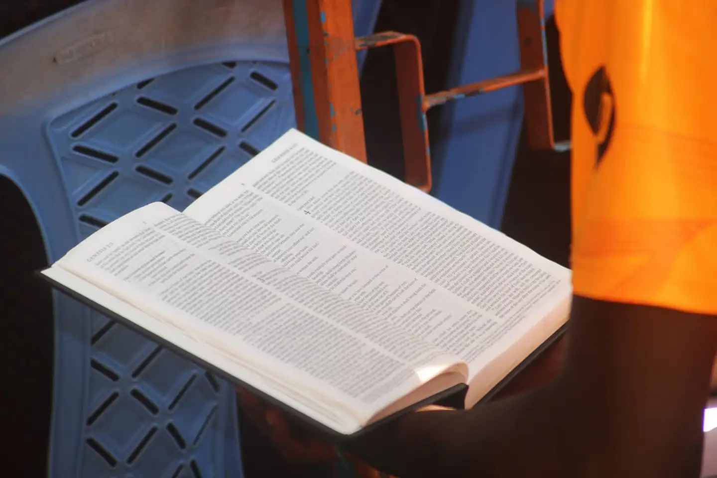 An open Bible held in someone’s hands beside a blue plastic chair