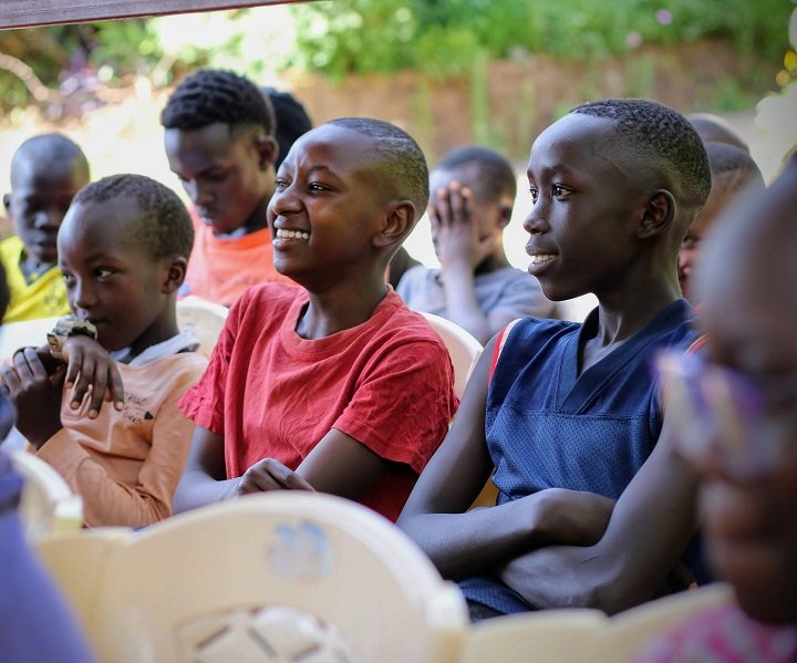 Children sitting together on plastic chairs outdoors, listening and smiling