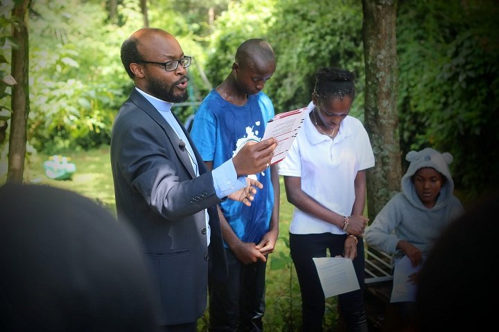A man reading aloud from printed sheets with three young people standing beside him in the garden